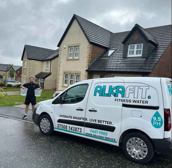 Gavin from AlkaFIT holding two bottles of alkaline water beside a branded AlkaFIT delivery van on a Lancashire street